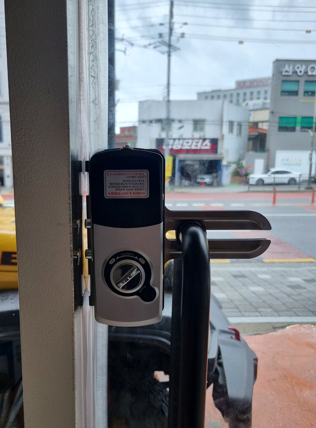 용산구 청파동 출장 — 유리문 틀에 물린 은색과 검은색 도어락, 돌림 손잡이와 막대 손잡이, 창밖 도로 풍경